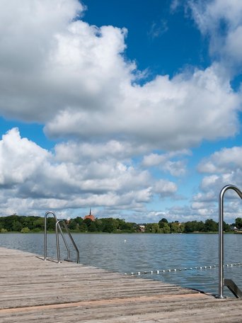 Badestellen & Bäder (© MHT.SH & Peter Lühr) Ein Holzsteg führt in den Bordesholmer See im Hintergrund sieht man eine Kirche. Der Himmel ist blau mit Wolken.