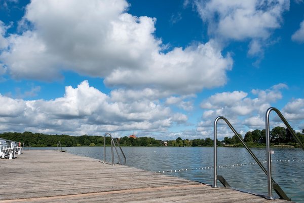 Badestellen & Bäder (© MHT.SH & Peter Lühr) Ein Holzsteg führt in den Bordesholmer See im Hintergrund sieht man eine Kirche. Der Himmel ist blau mit Wolken.